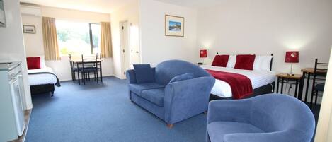 A hotel room with a bed, two blue armchairs, a desk, and a chair. There is a window with curtains and a picture on the wall.