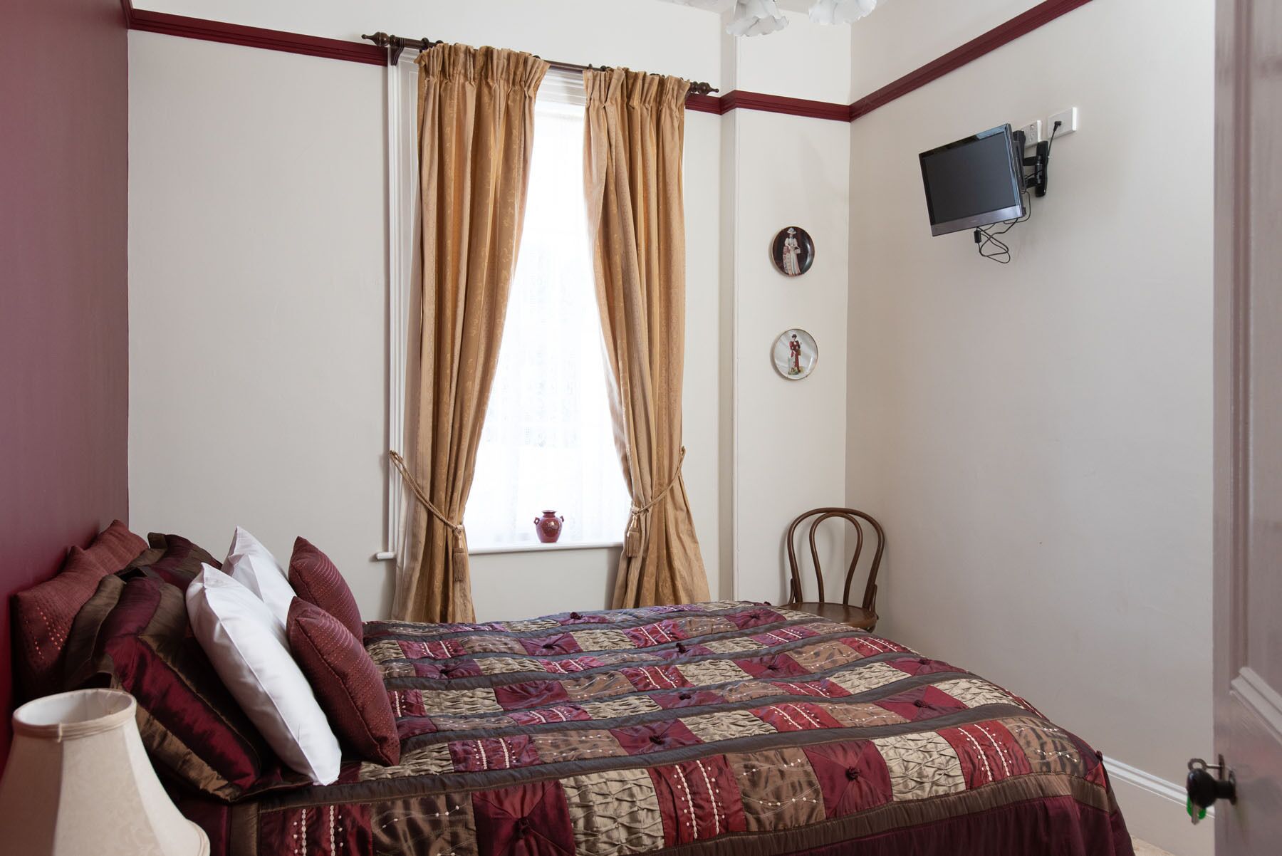 Ein Schlafzimmer mit Bett, einem Fenster mit Vorhängen, einem an der Wand montierten Fernseher und einem kleinen Tisch mit Lampe.