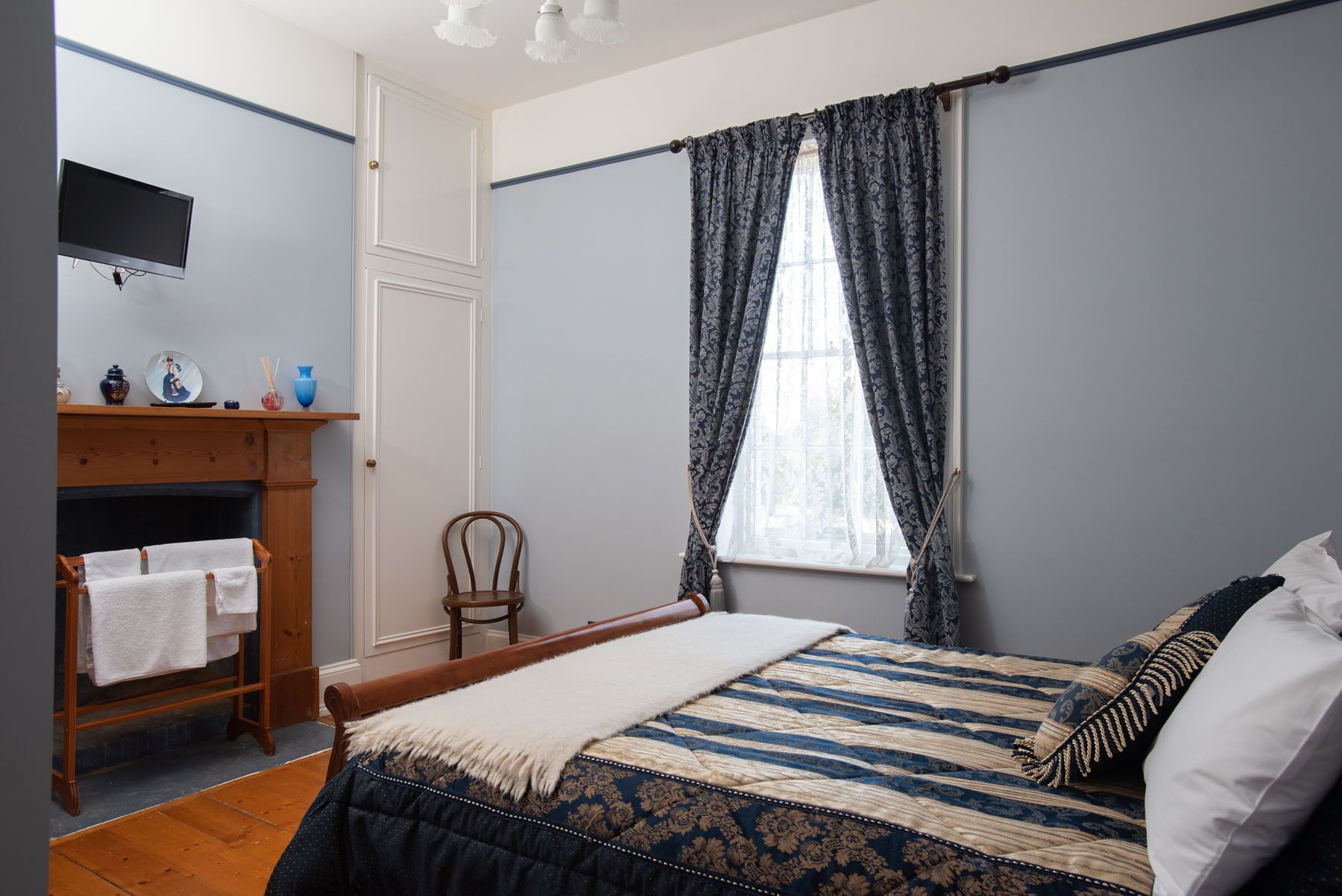 A bedroom with a bed, a wooden chair, a small table with decorative items, and a window with curtains.