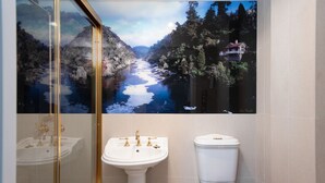 A bathroom with a large scenic mural of a river and forest, a white pedestal sink with gold fixtures, and a white toilet.