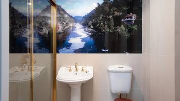 A bathroom with a large scenic mural of a river and forest, a white pedestal sink with gold fixtures, and a white toilet.