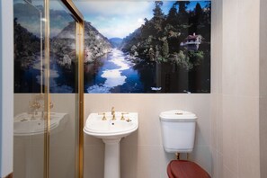 A bathroom with a large scenic mural of a river and forest, a white pedestal sink with gold fixtures, and a white toilet.