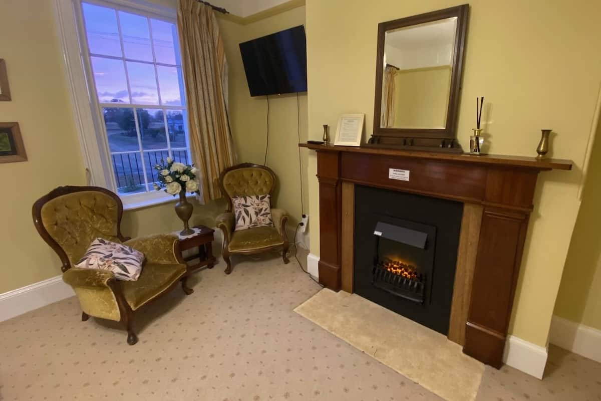 Una habitación con chimenea, dos sillones, una mesita con un florero, un televisor montado en la pared y una ventana con cortinas.
