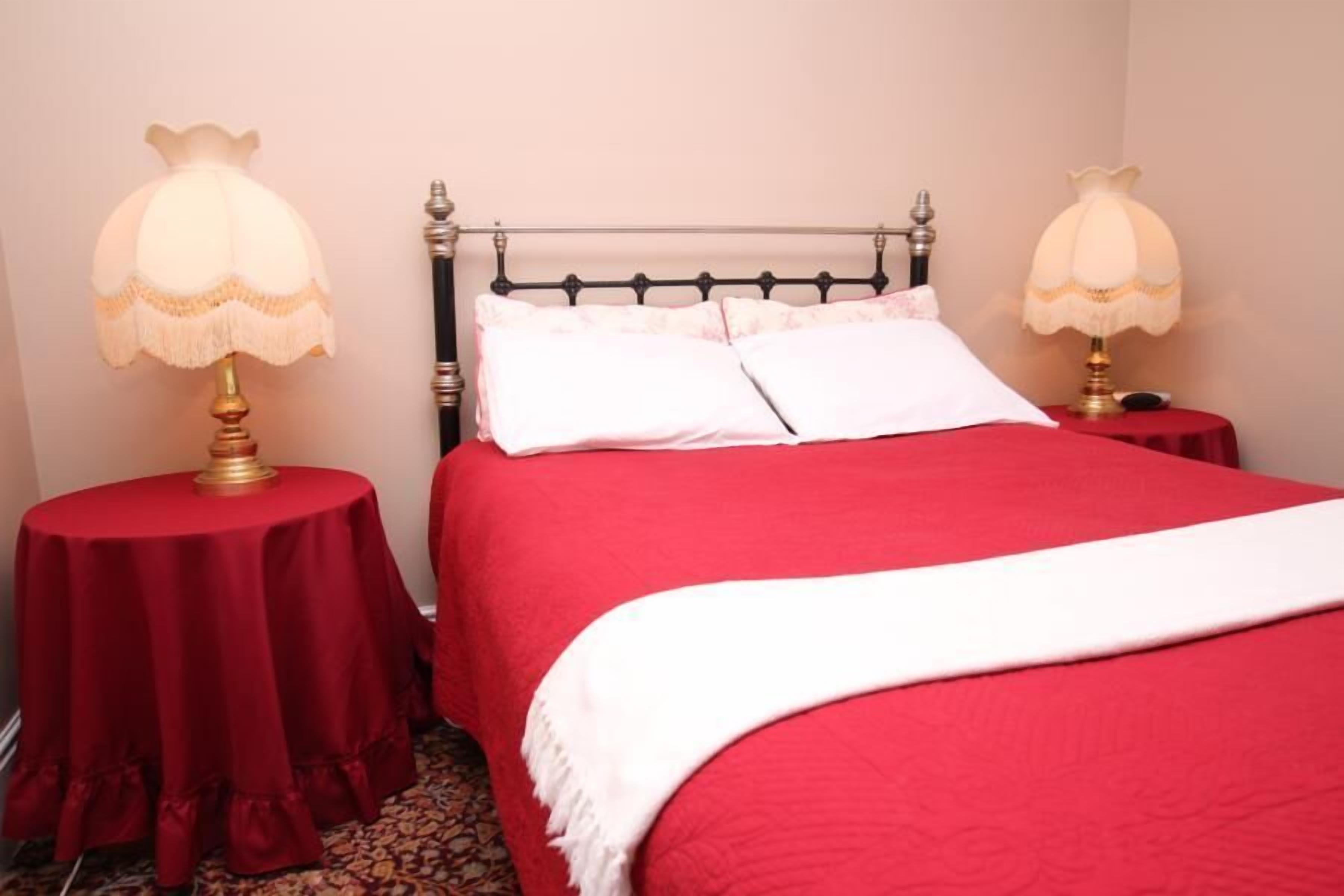 A neatly made bed with a red bedspread and white pillows, flanked by two round tables with lamps, in a room with a simple, clean design.