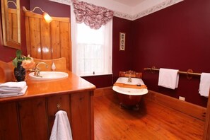 Une salle de bain avec un meuble-lavabo en bois, une baignoire sur pieds et une fenêtre agrémentée d’un rideau à motifs floraux.