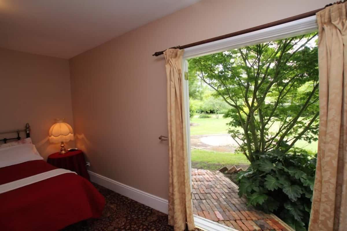 Habitación de hotel con cama, lámpara de noche y vista a un jardín por una puerta abierta.