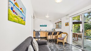 A modern living room with a grey sofa, a dining area with a table and chairs, a kitchen with a stainless steel sink, and a large glass door leading to an outdoor deck. - Waipu Cove Resort (Waipu)