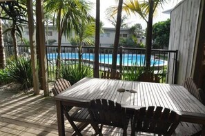 A wooden deck with a table and chairs, overlooking a swimming pool surrounded by palm trees. - Waipu Cove Resort (Waipu)