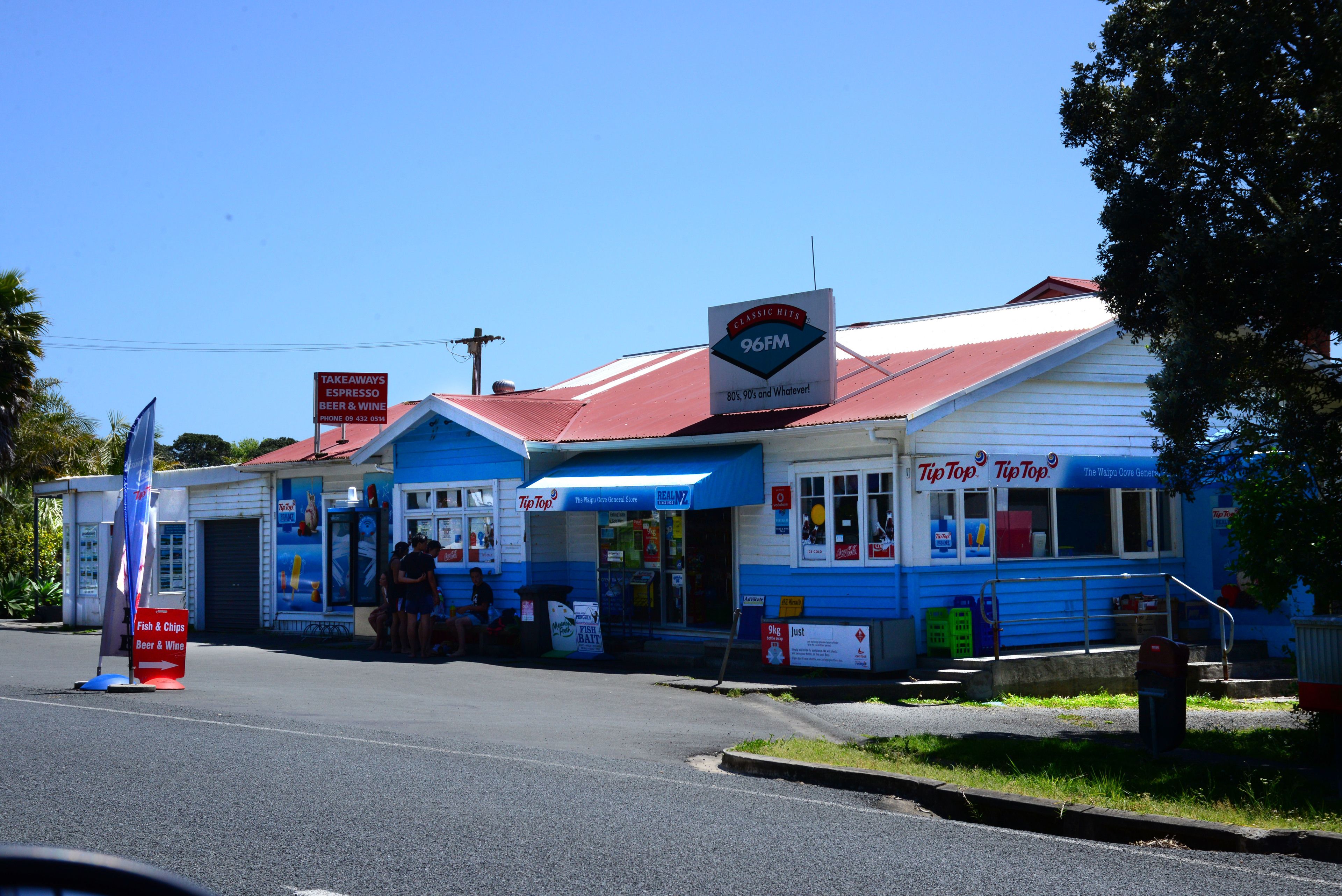 A small, two-story building with a red roof and blue awnings. There are signs for a radio station and a place offering fish & chips, beer, and wine.
