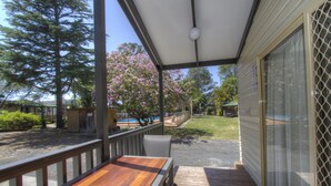 A covered outdoor deck with a wooden table and chairs, a view of a swimming pool, and a lawn with trees and a fence. - Twin Dolphins Holiday Park (Tuncurry)
