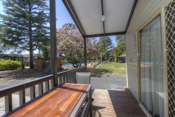A covered outdoor deck with a wooden table and chairs, a view of a swimming pool, and a lawn with trees and a fence. - Twin Dolphins Holiday Park (Tuncurry)