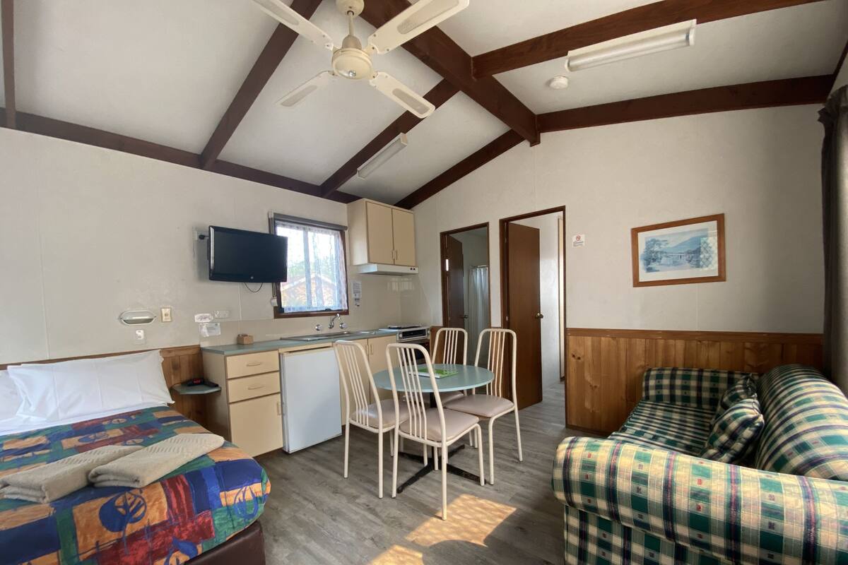 A compact room with a bed, a small kitchenette, a dining table with chairs, a sofa, and a ceiling fan.