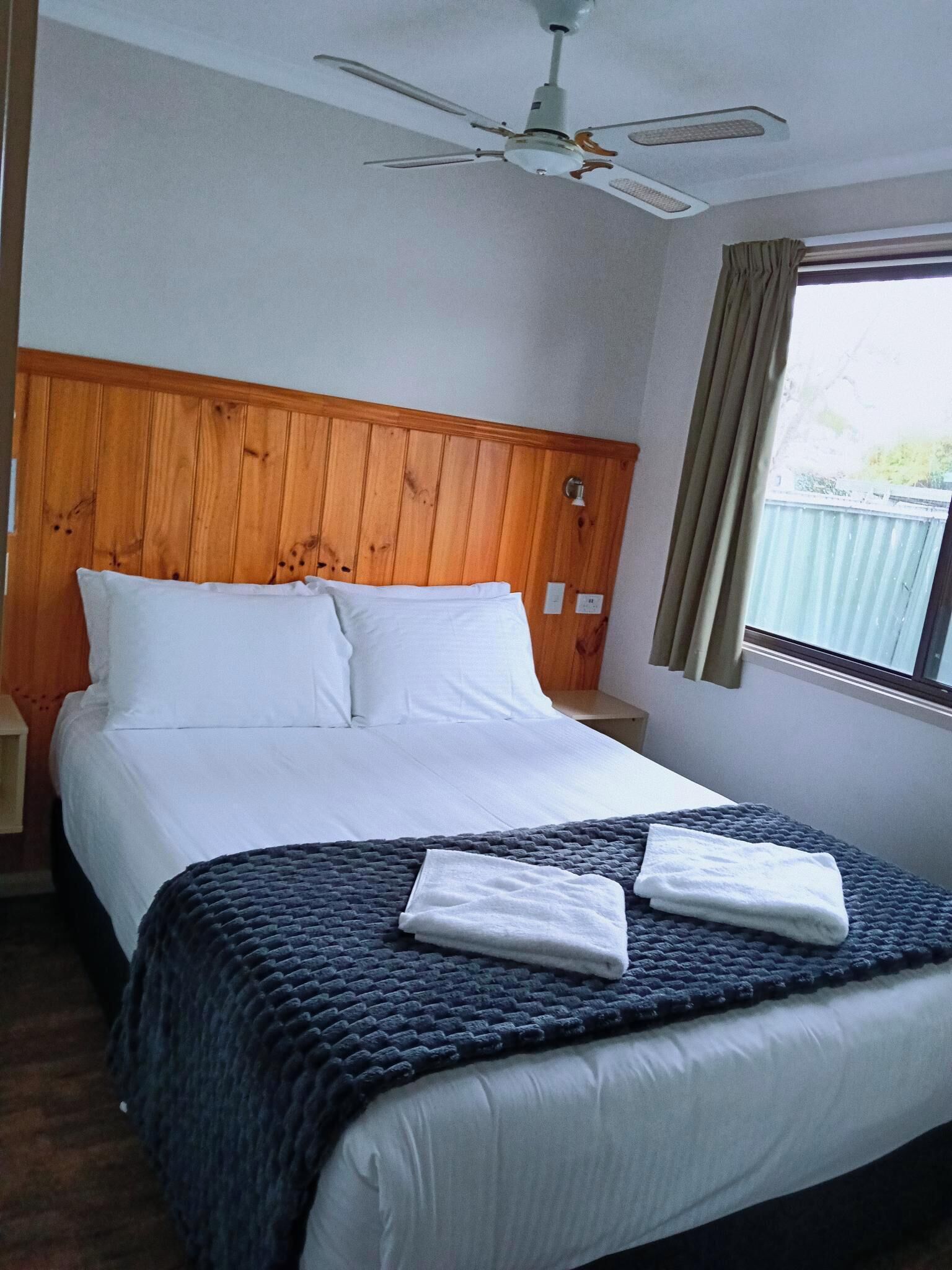 A hotel room with a wooden headboard, a bed with white linens, a dark blue blanket, and a ceiling fan.
