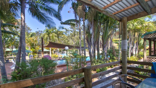 A wooden deck with a glass table and wicker chairs, overlooking a pool area with palm trees and a covered seating area. - Twin Dolphins Holiday Park (Tuncurry)