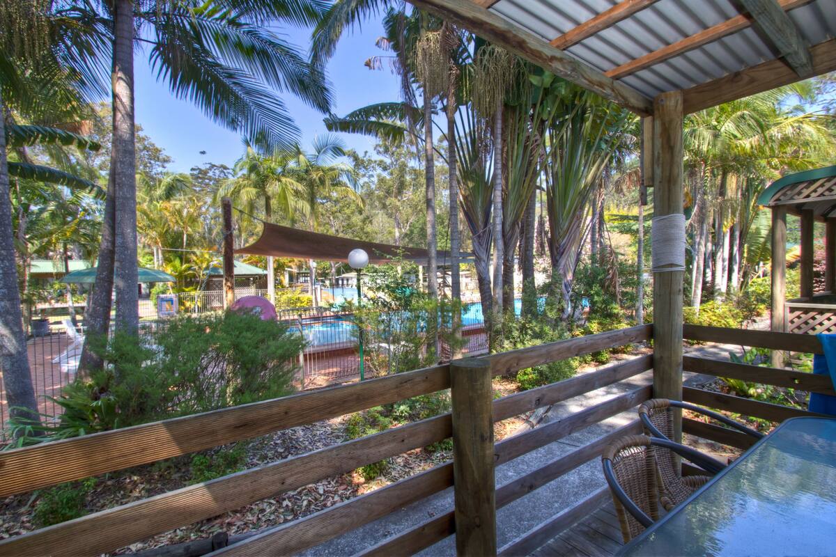 A wooden deck with a glass table and wicker chairs, overlooking a pool area with palm trees and a covered seating area.