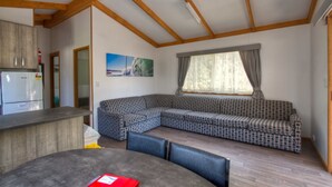 A modern living room with a wooden ceiling, a built-in kitchen area, a large sectional sofa, and a dining table with chairs. - Twin Dolphins Holiday Park (Tuncurry)