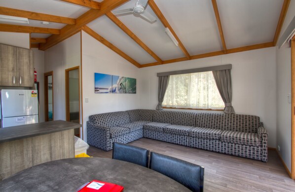 A modern living room with a wooden ceiling, a built-in kitchen area, a large sectional sofa, and a dining table with chairs. - Twin Dolphins Holiday Park (Tuncurry)