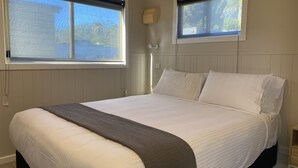 Ein ordentlich bezogenes Bett mit weißen Laken und einer grauen Tagesdecke in einem Zimmer mit Holzvertäfelung und zwei Fenstern.