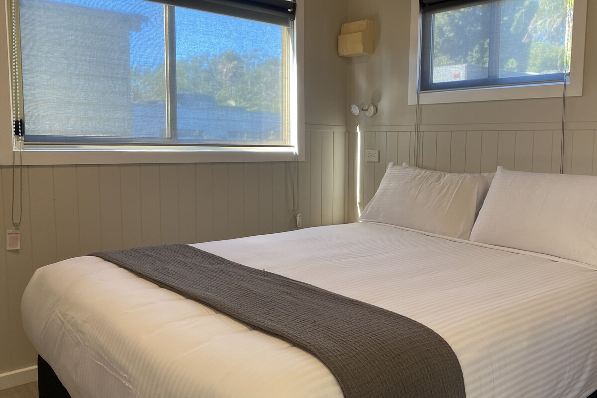 A neatly made bed with white linens and a gray throw blanket in a room with wooden paneling and two windows.