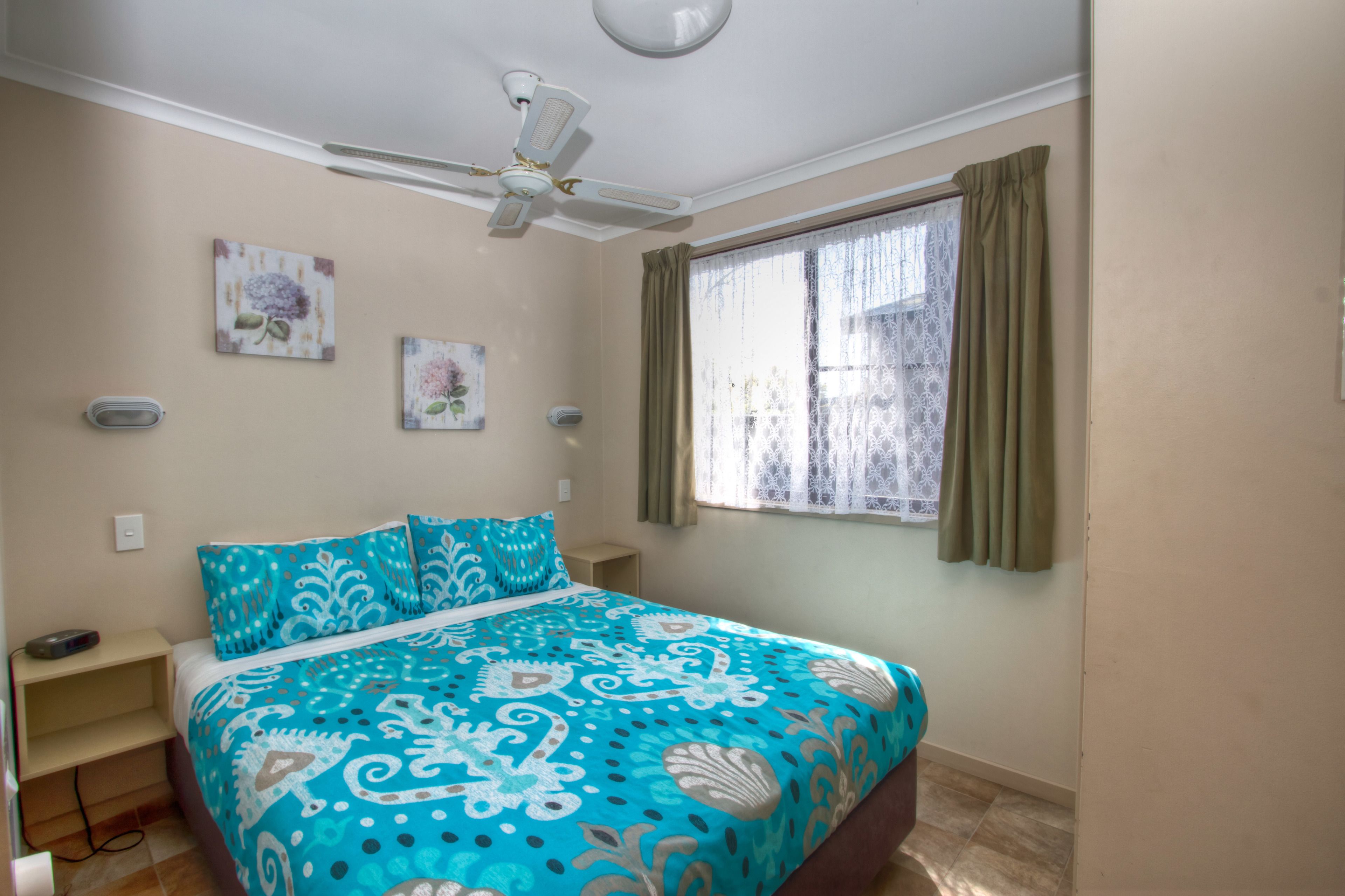 Un dormitorio con cama, ventilador de techo, ventana con cortinas y cuadros en la pared.