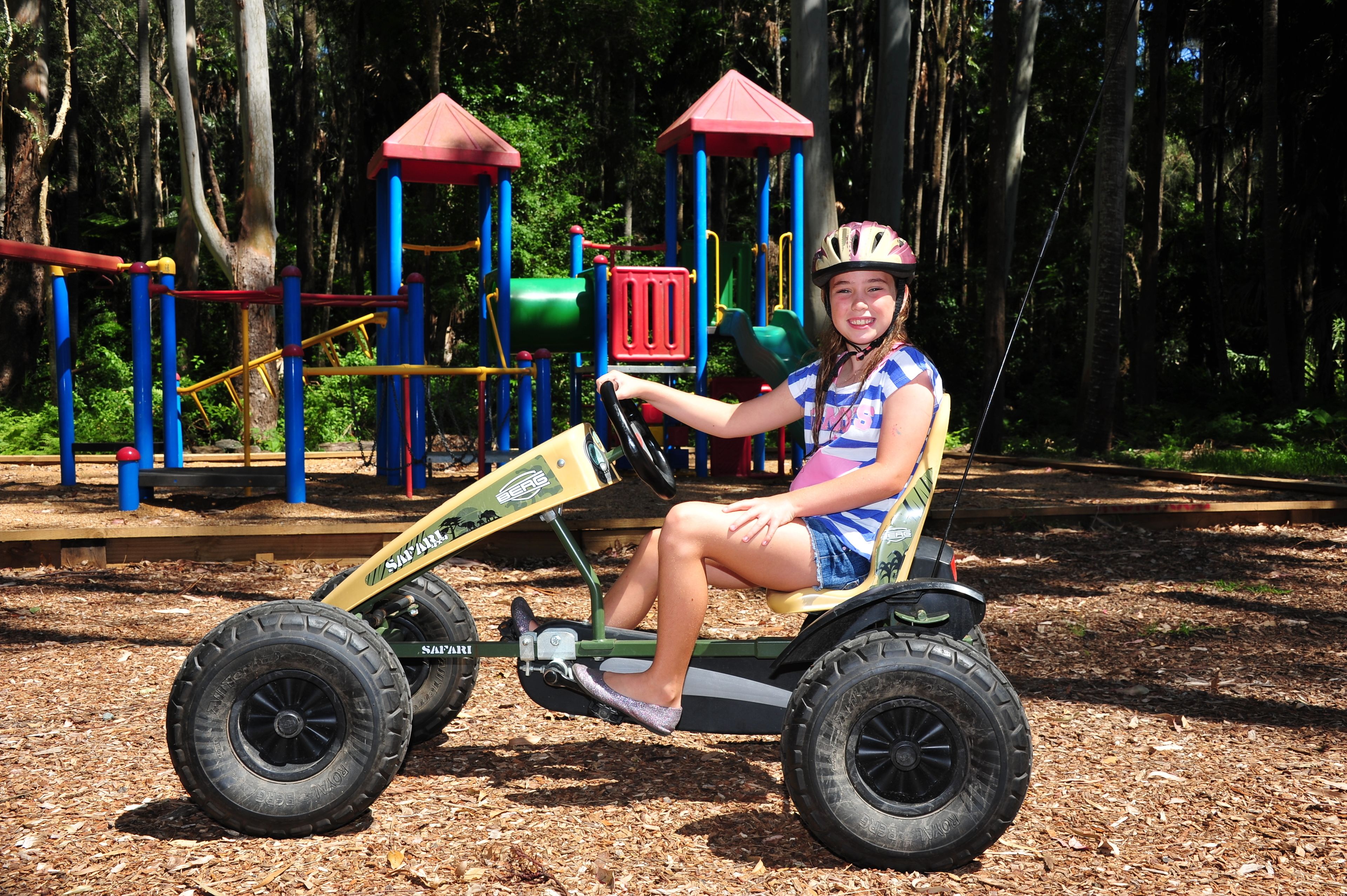 Un enfant est assis sur un petit tracteur à pédales dans un parc, avec des jeux et des arbres en arrière-plan.