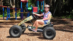 A child on a ride-on toy tractor at a playground with play structures and trees in the background. - Twin Dolphins Holiday Park (Tuncurry)