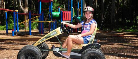 Un enfant est assis sur un petit tracteur à pédales dans un parc, avec des jeux et des arbres en arrière-plan.