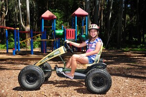 A child on a ride-on toy tractor at a playground with play structures and trees in the background. - Twin Dolphins Holiday Park (Tuncurry)