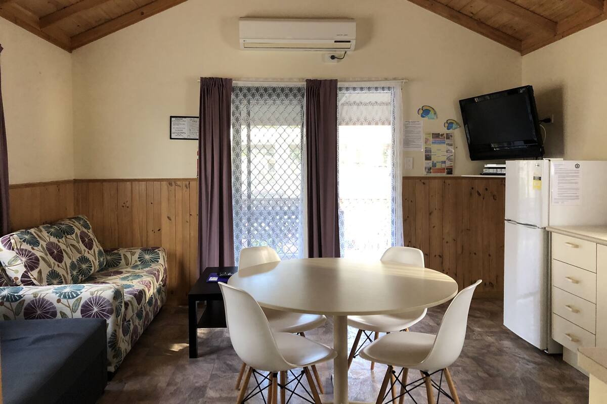 A cozy room with a wooden ceiling, a white table with four chairs, a sofa with a floral pattern, a refrigerator, and a television on the wall.