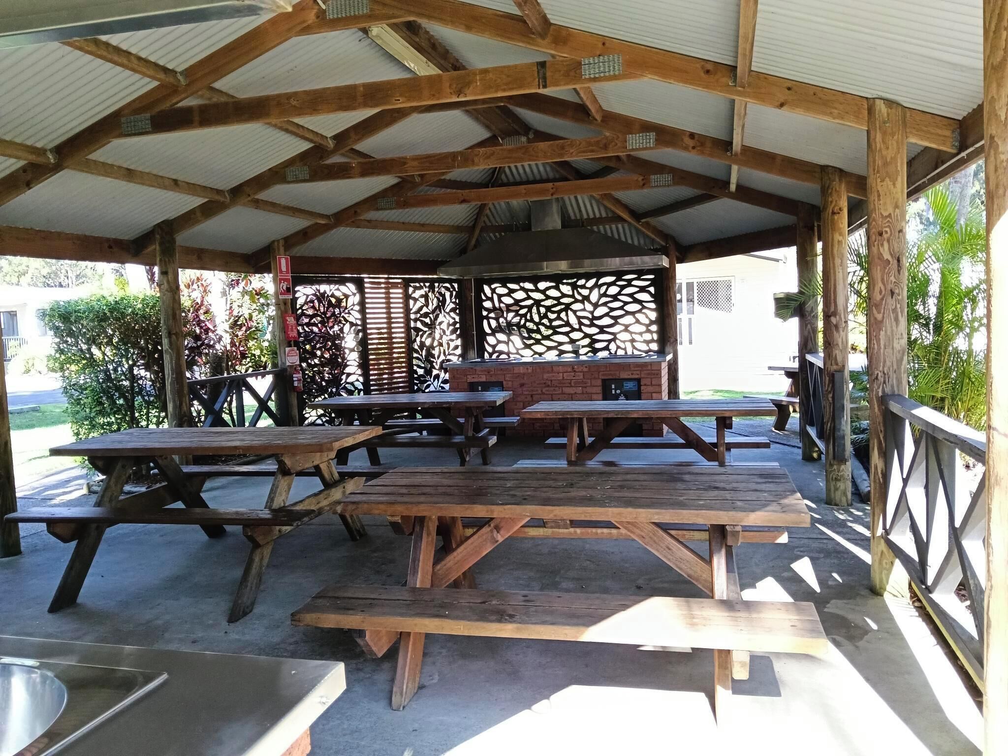 An outdoor covered picnic area with multiple wooden picnic tables and benches, a brick barbecue grill, and a metal sink.