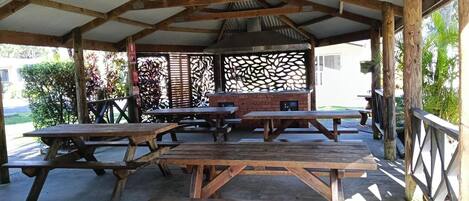 An outdoor covered picnic area with multiple wooden picnic tables and benches, a brick barbecue grill, and a metal sink.