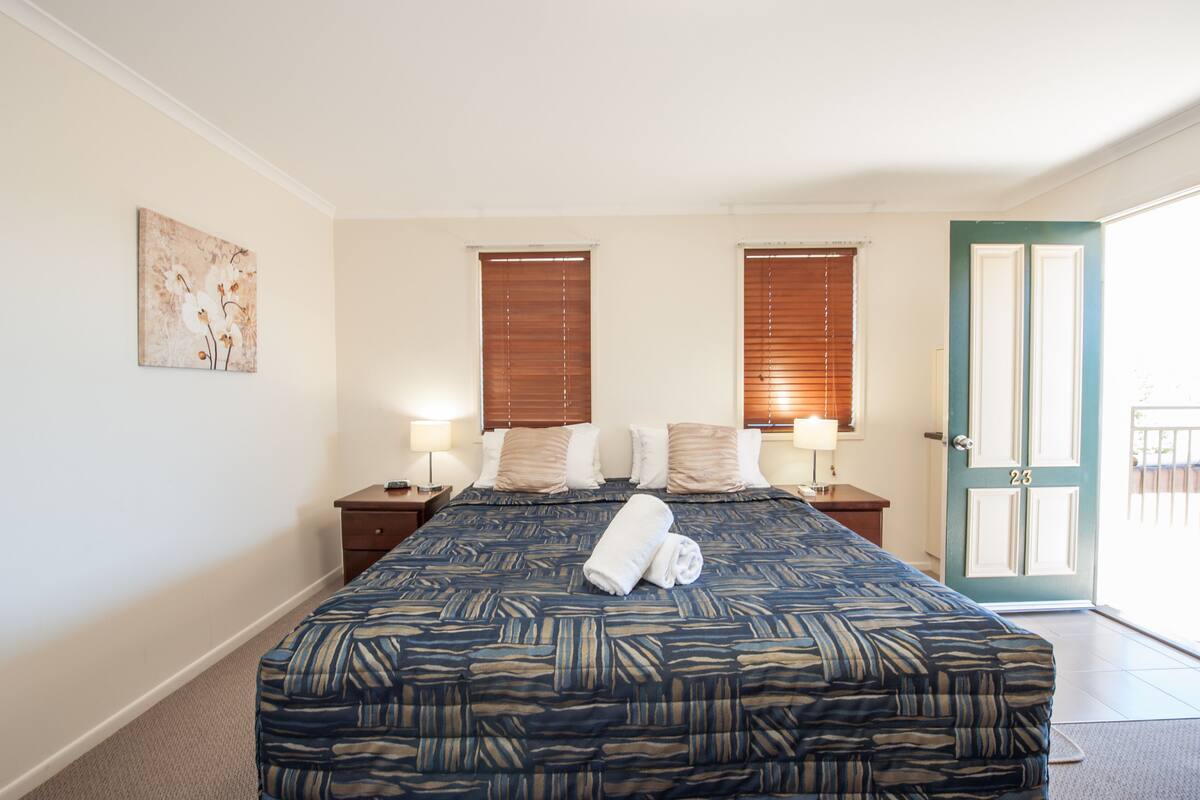 A hotel room with a large bed, two bedside tables with lamps, a door with a number, and a window with blinds.