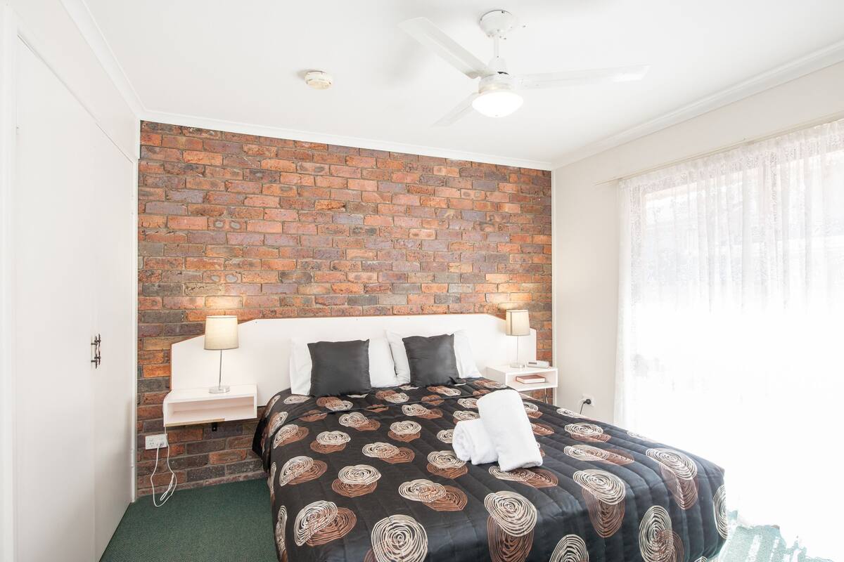 A hotel room with a brick-patterned wall, a bed with a patterned bedspread, two bedside tables with lamps, and a ceiling fan.