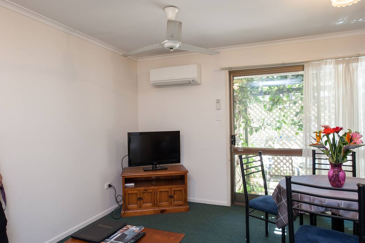 A living room with a flat-screen TV on a wooden stand, a ceiling fan, and a dining table with chairs.