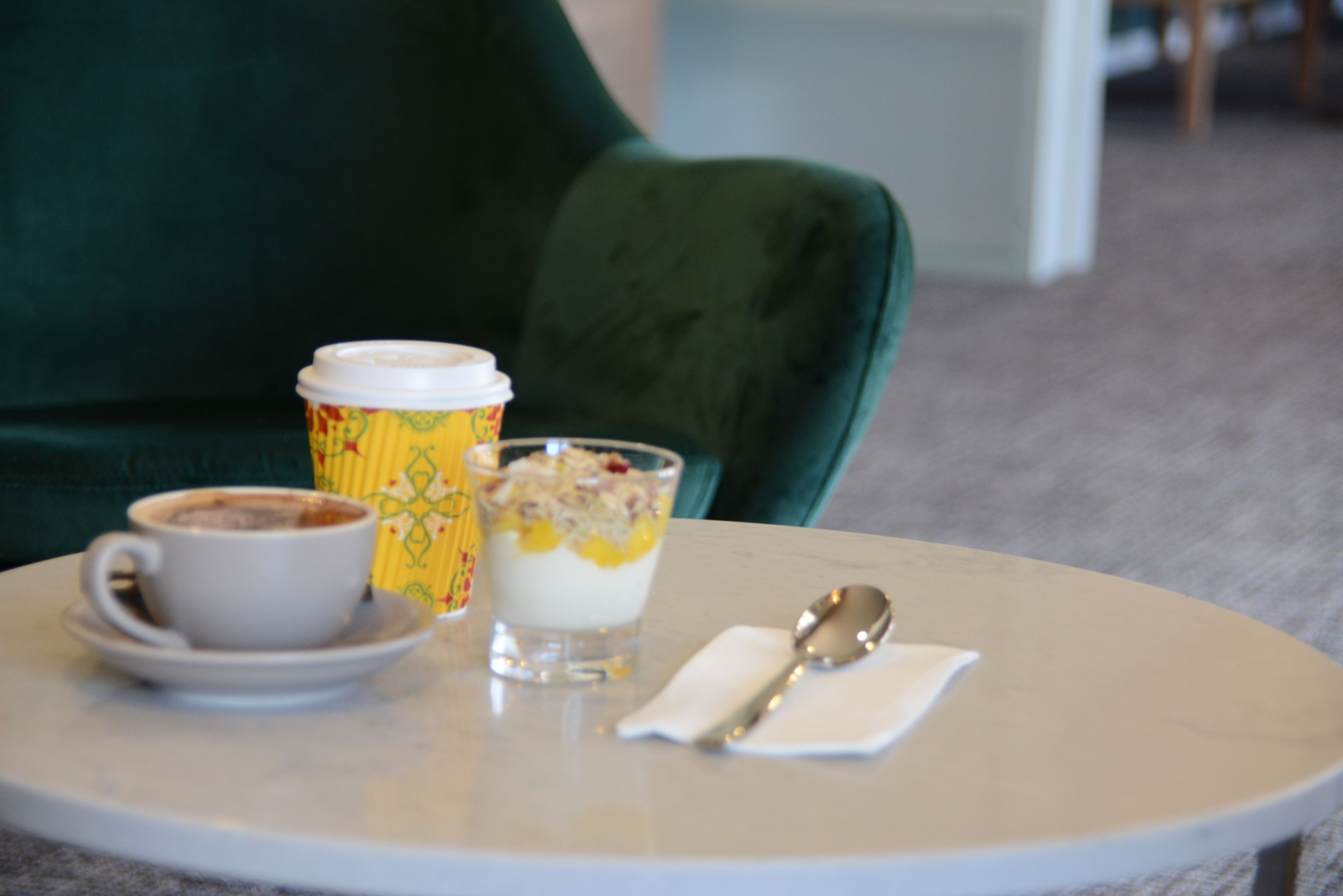 A table with a cup of coffee, a glass of yogurt with fruit, and a spoon on a white tablecloth.