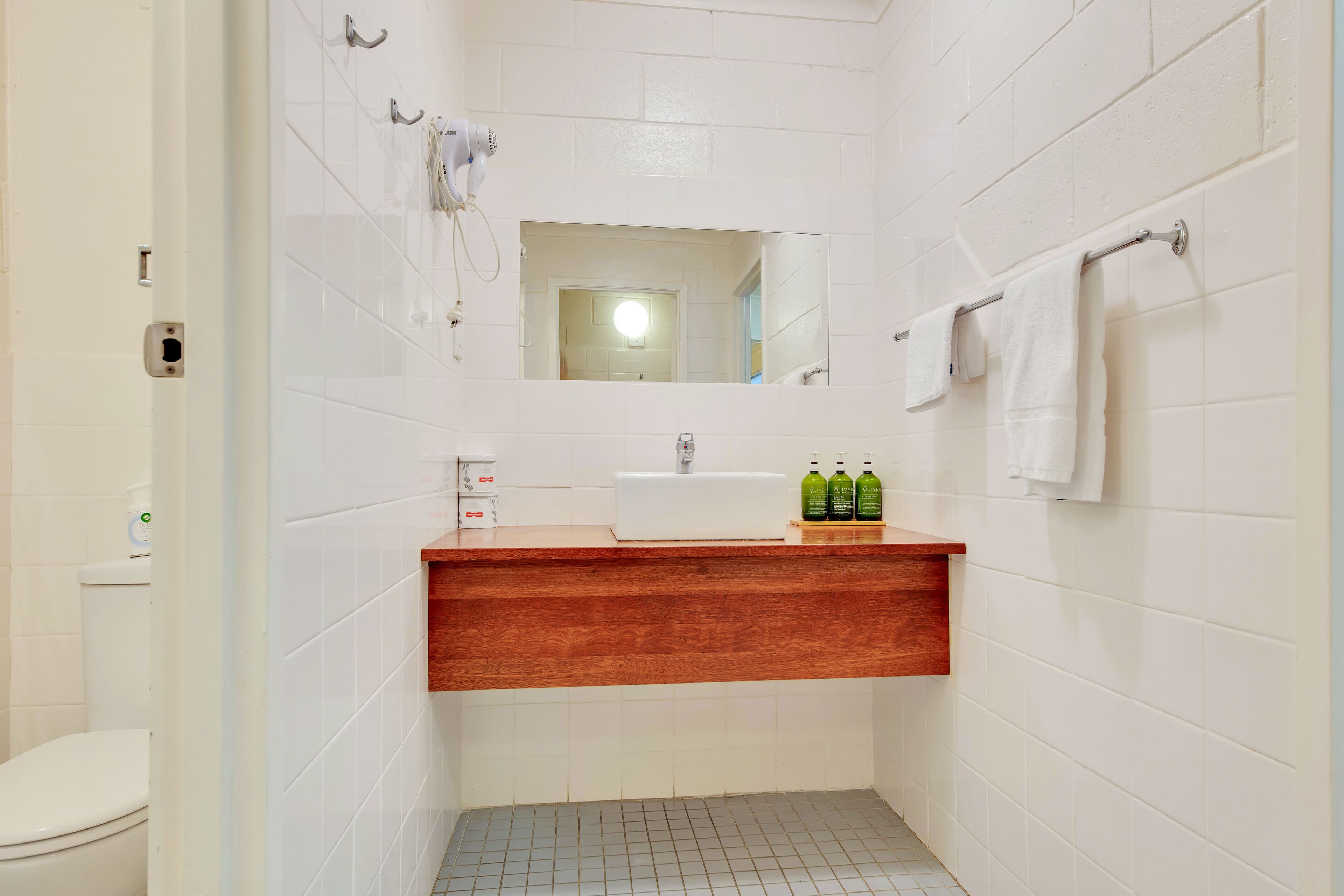 Un baño con azulejos blancos, un tocador de madera, un espejo, un perchero para toallas y un inodoro.