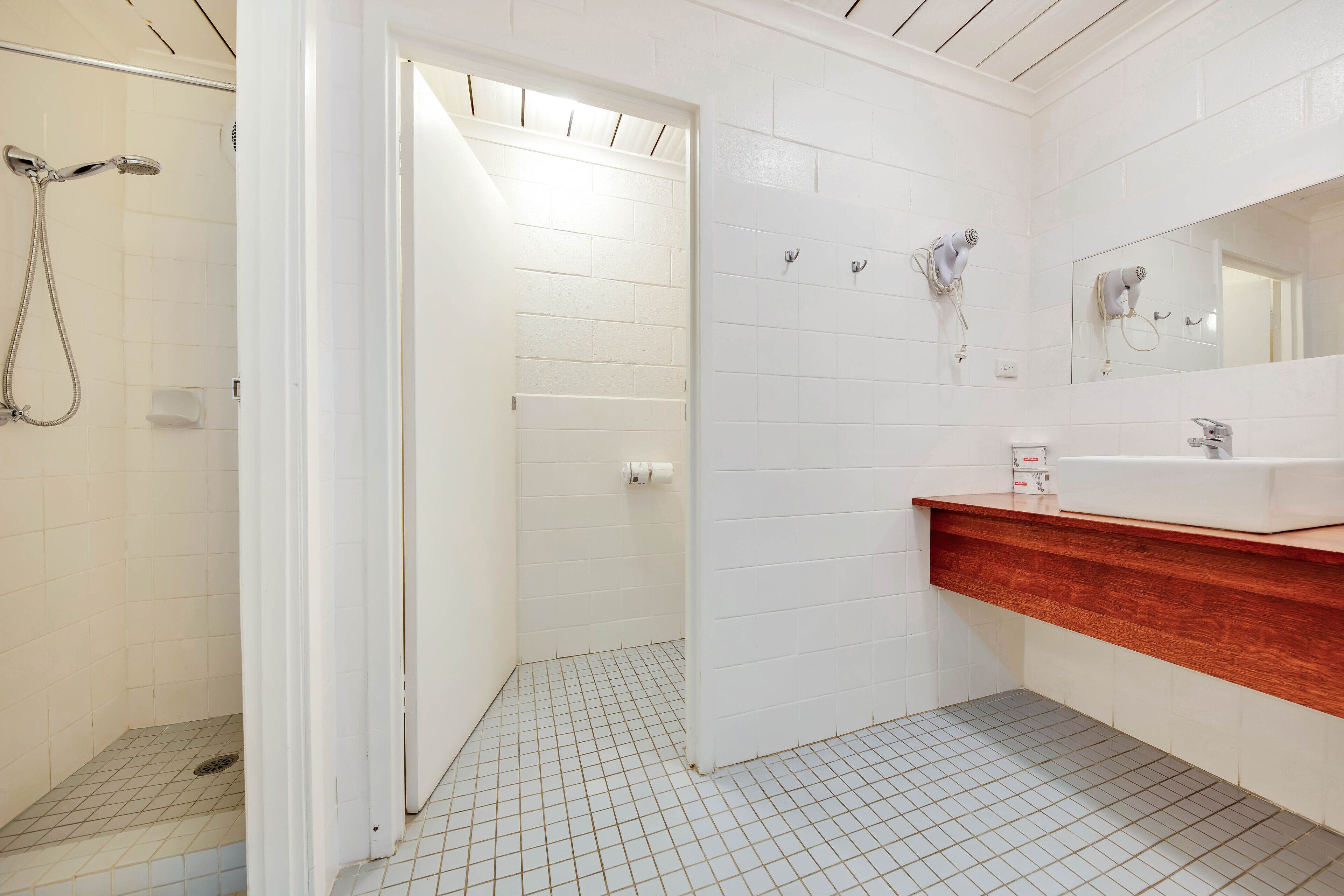 Un baño con paredes de azulejos blancos, zona de ducha, tocador de madera y un espejo.