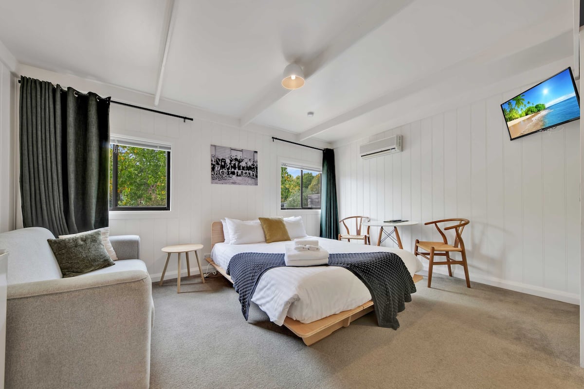 A bedroom with a bed, a sofa, a small table, a chair, and a TV on the wall.