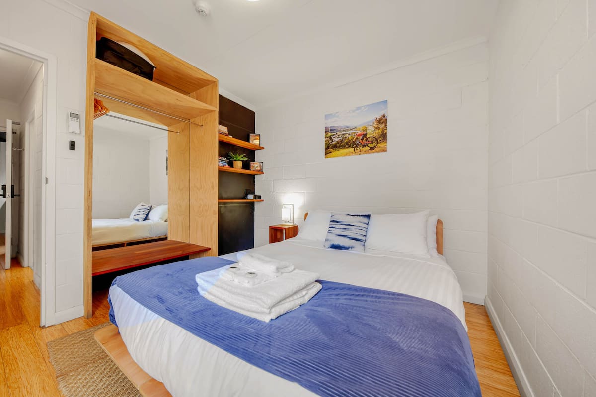 A bedroom with a bed, a wooden wardrobe, a shelf with books and a plant, a wall-mounted picture, and a bedside table with a lamp.