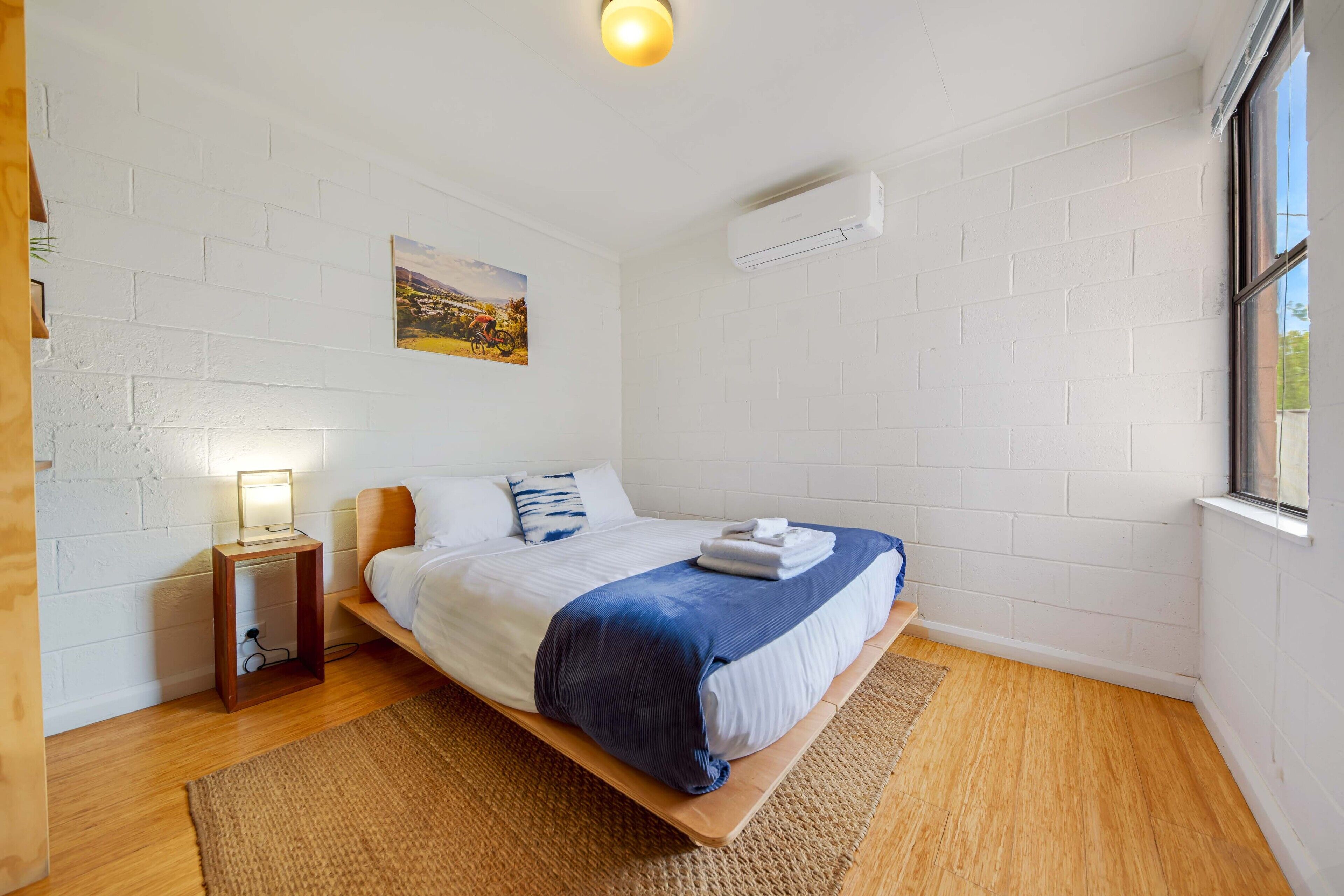 A bedroom with a bed, a bedside table, a lamp, a picture on the wall, and an air conditioning unit.