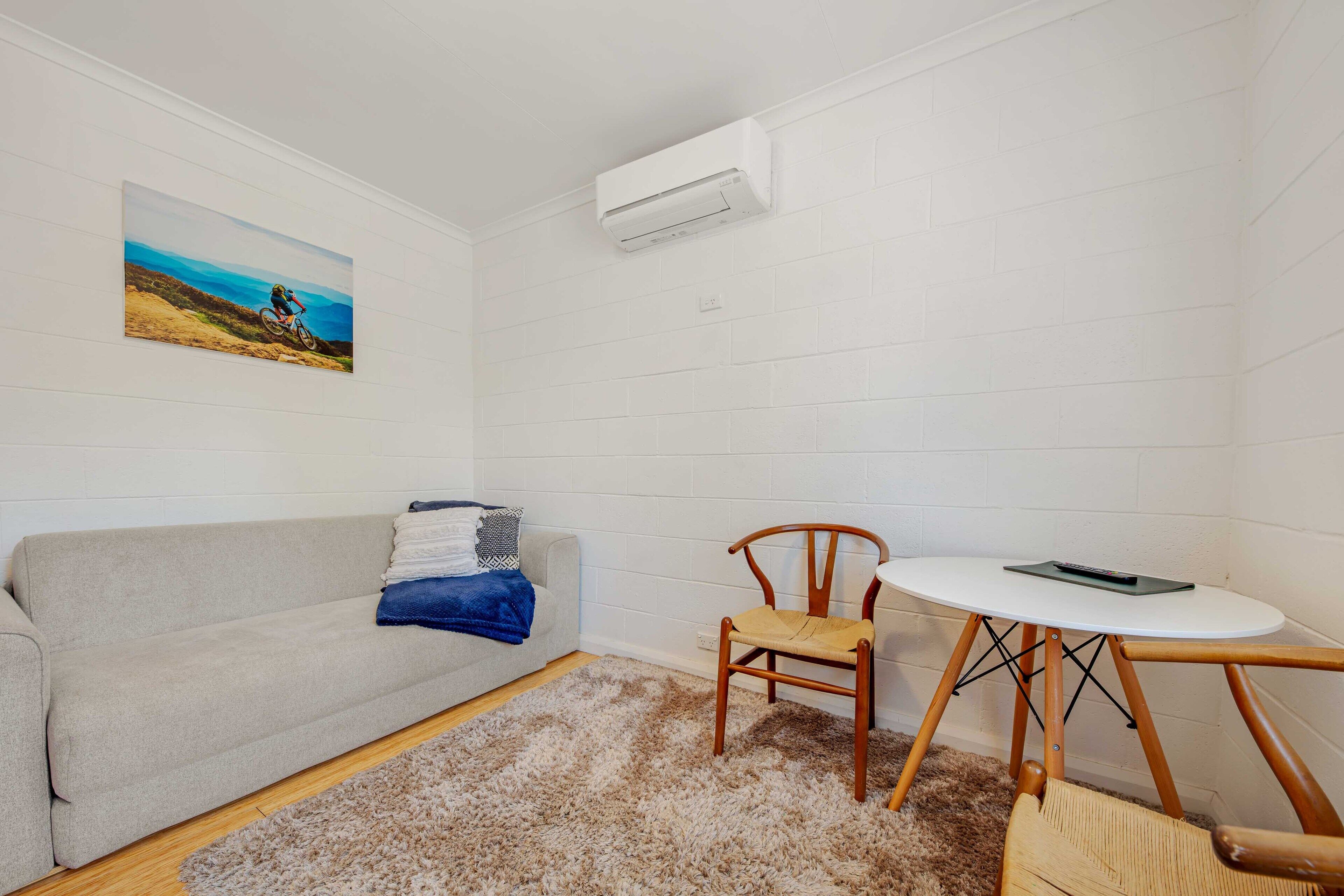 A room with a white brick wall, a grey sofa, a small round table with a chair, and a mounted air conditioner.