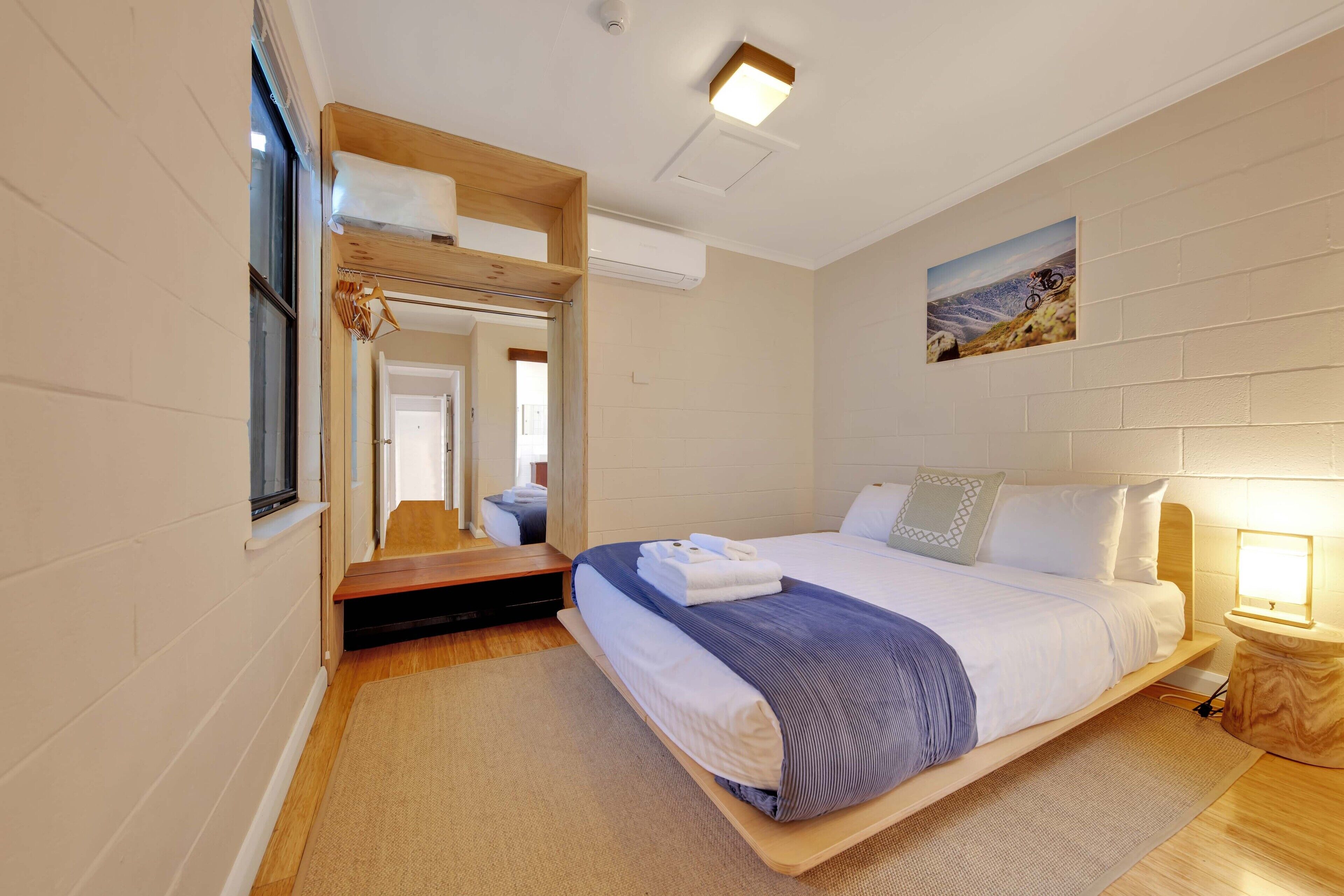 A modern hotel room with a large bed, a wooden headboard, a small bedside table with a lamp, a mirror, and a window with a view of the mountains.