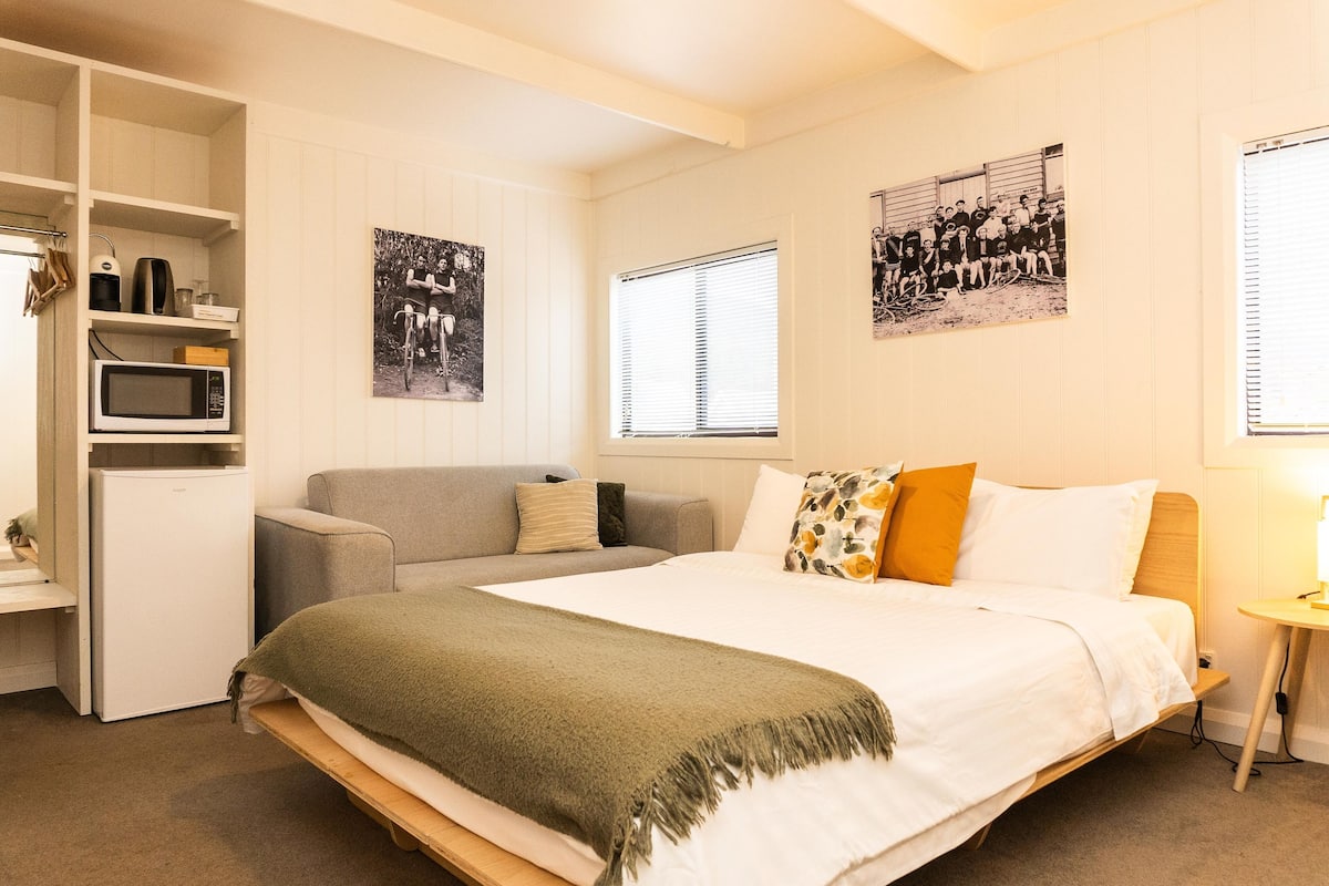 A bedroom with a bed, a sofa, a small kitchenette, and a window with blinds.