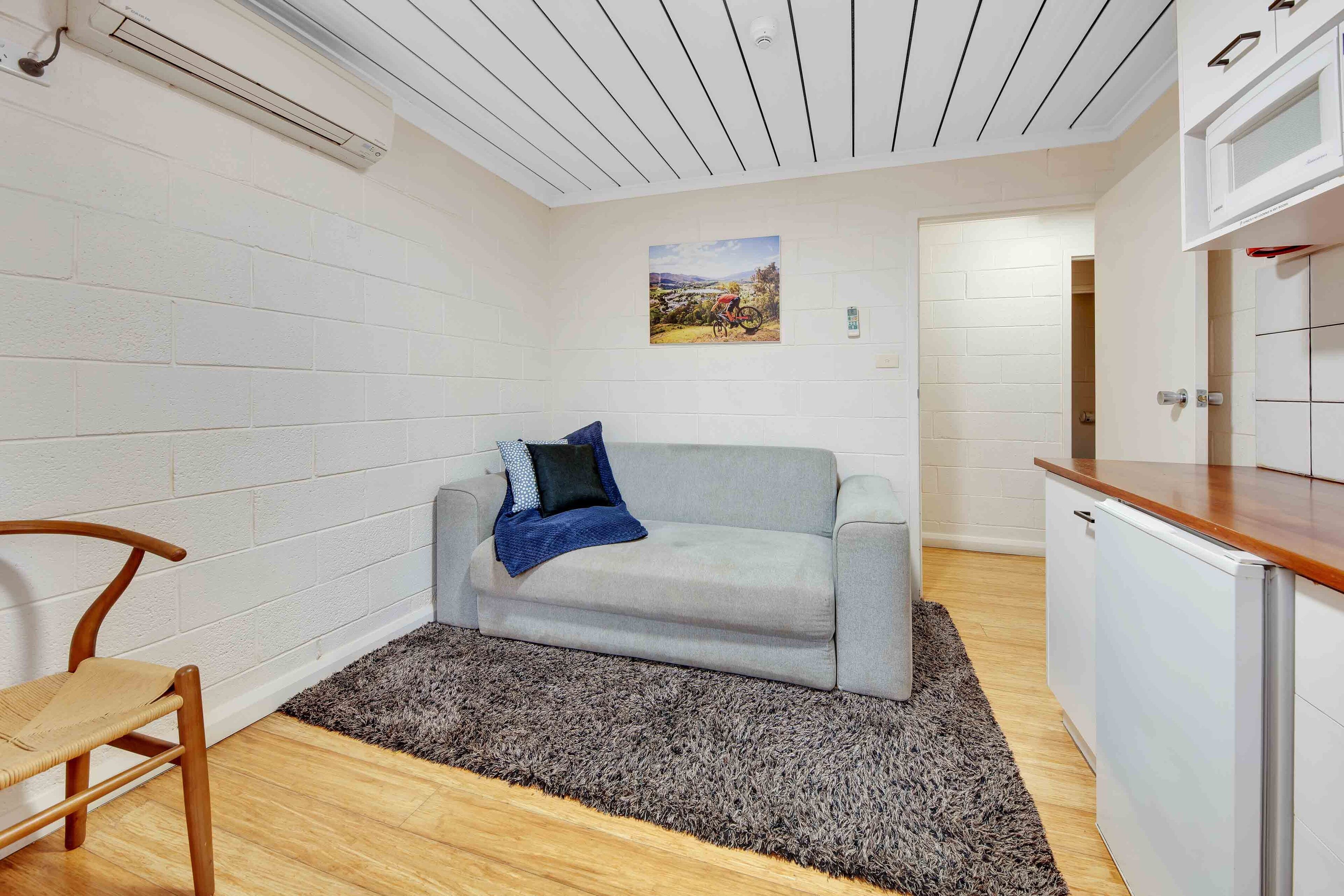 A compact living space with a grey sofa, a wooden chair, a kitchen area with a white refrigerator, and a grey shag rug.
