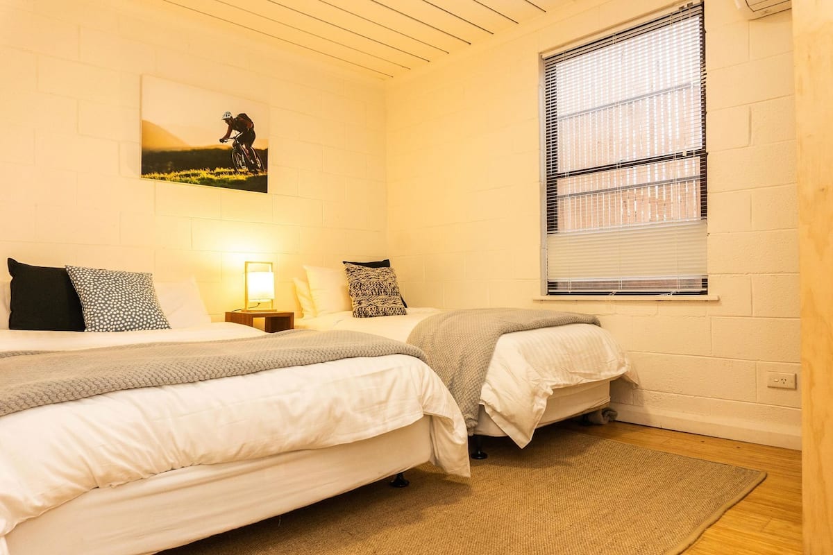 A bedroom with two beds, a window with blinds, a wall-mounted picture, and a bedside lamp.