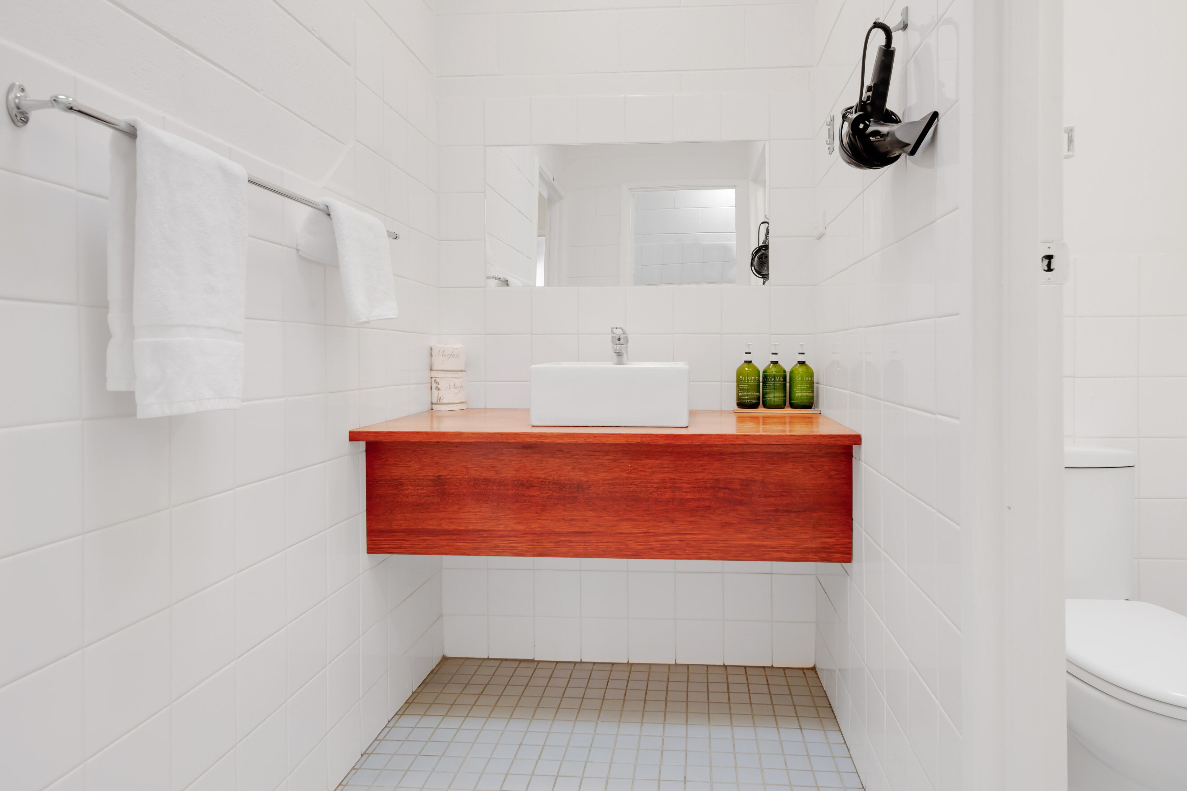 A bathroom with white tiled walls, a wooden vanity, a white sink, and a mirror above it. There are two towels hanging on a towel rack, and a green bottle on the vanity.