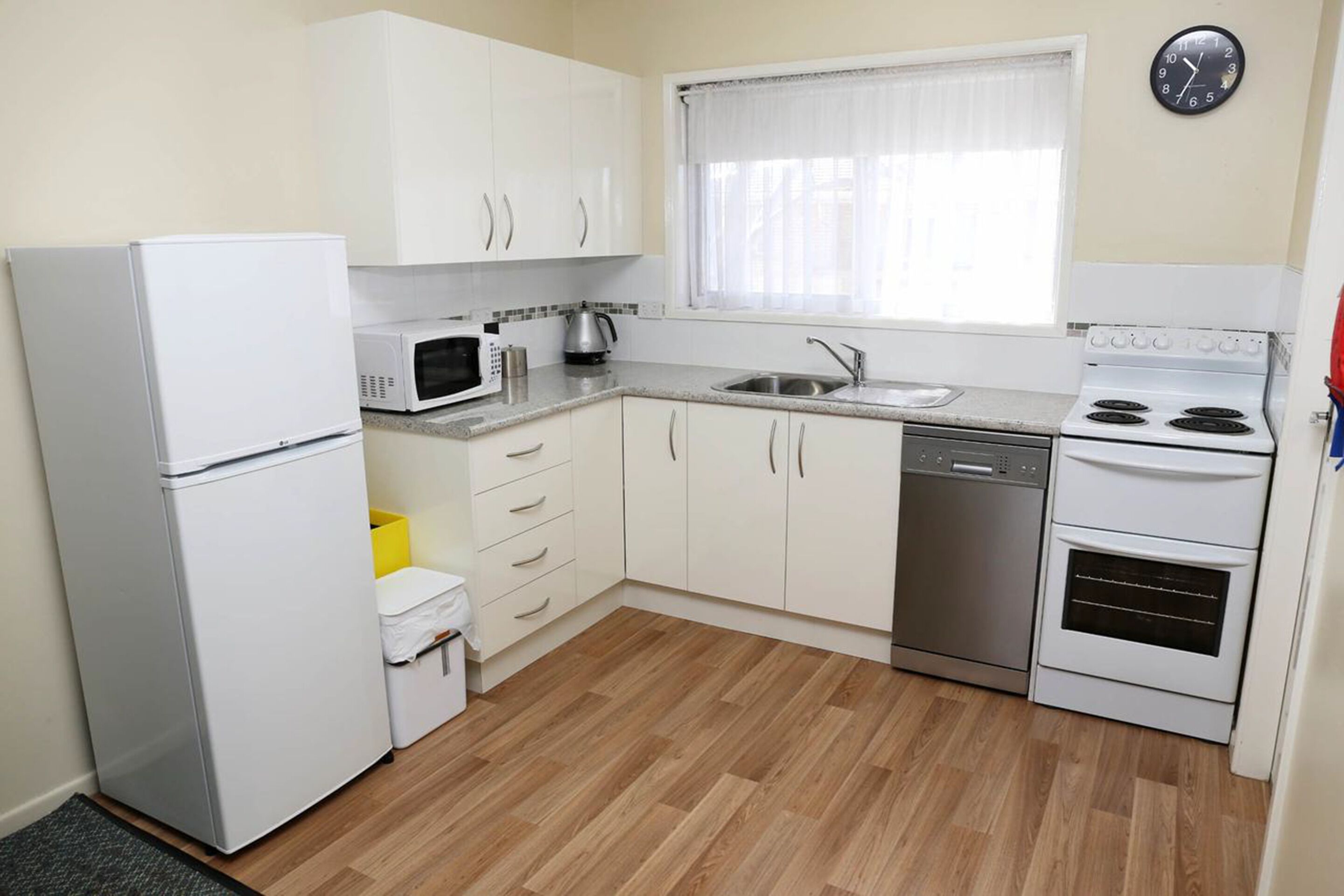 Een kleine keuken met witte kastjes, een koelkast, een magnetron, een waterkoker, een spoelbak en een oven.