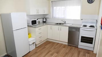 Een kleine keuken met witte kastjes, een koelkast, een magnetron, een waterkoker, een spoelbak en een oven.