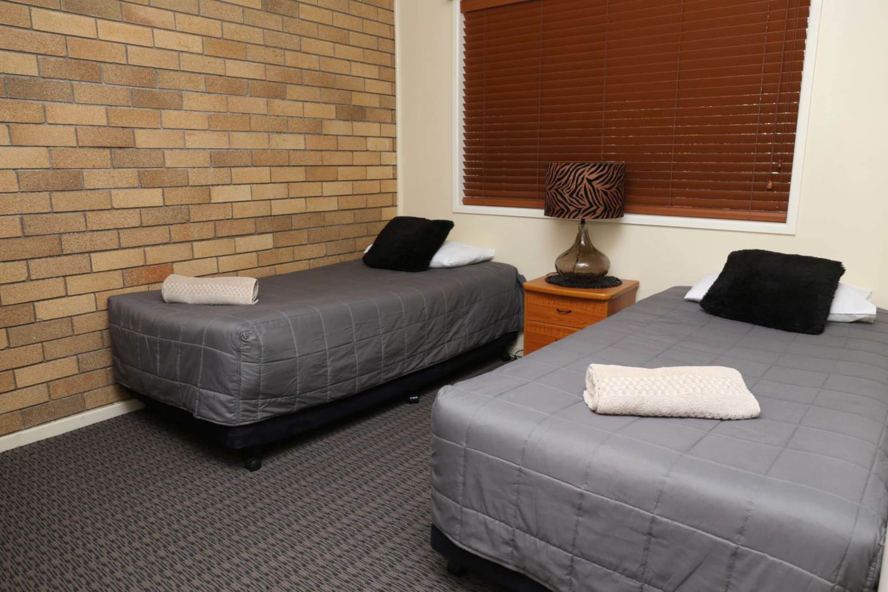 Two single beds with grey bedding, a wooden nightstand, a lamp, and a window with blinds.