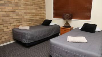 Two single beds with grey bedding, a wooden nightstand, a lamp, and a window with blinds.